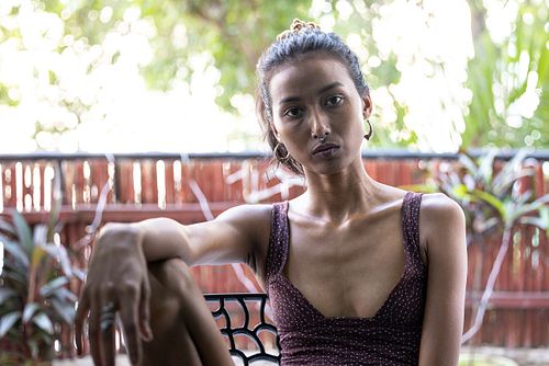 A slender Indian woman in a purple top is sitting on a chair on a balcony, with her arm placed on her knee.