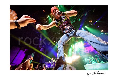 Horizontal color image of Bret Michaels performing live during a Poison concert, leaning toward the crowd with microphone in hand under vivid stage lights