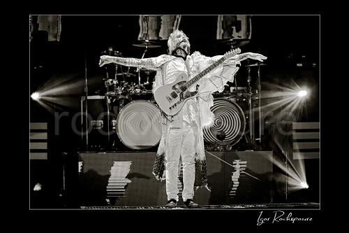 Horizontal black and white image of John 5 performing live on stage with an electric guitar, white stage outfit, outstretched arms, and bright backlights