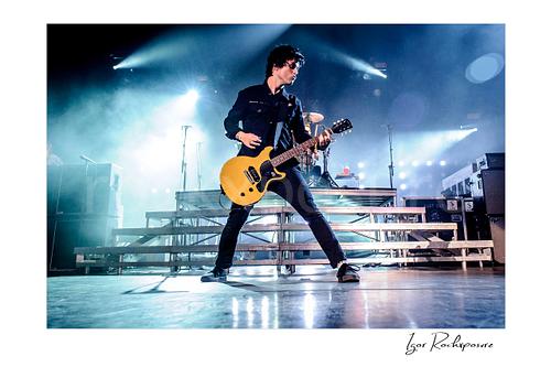 Horizontal color image of Billie Joe Armstrong of Green Day playing guitar onstage in a wide stance under bright concert lights and stage haze