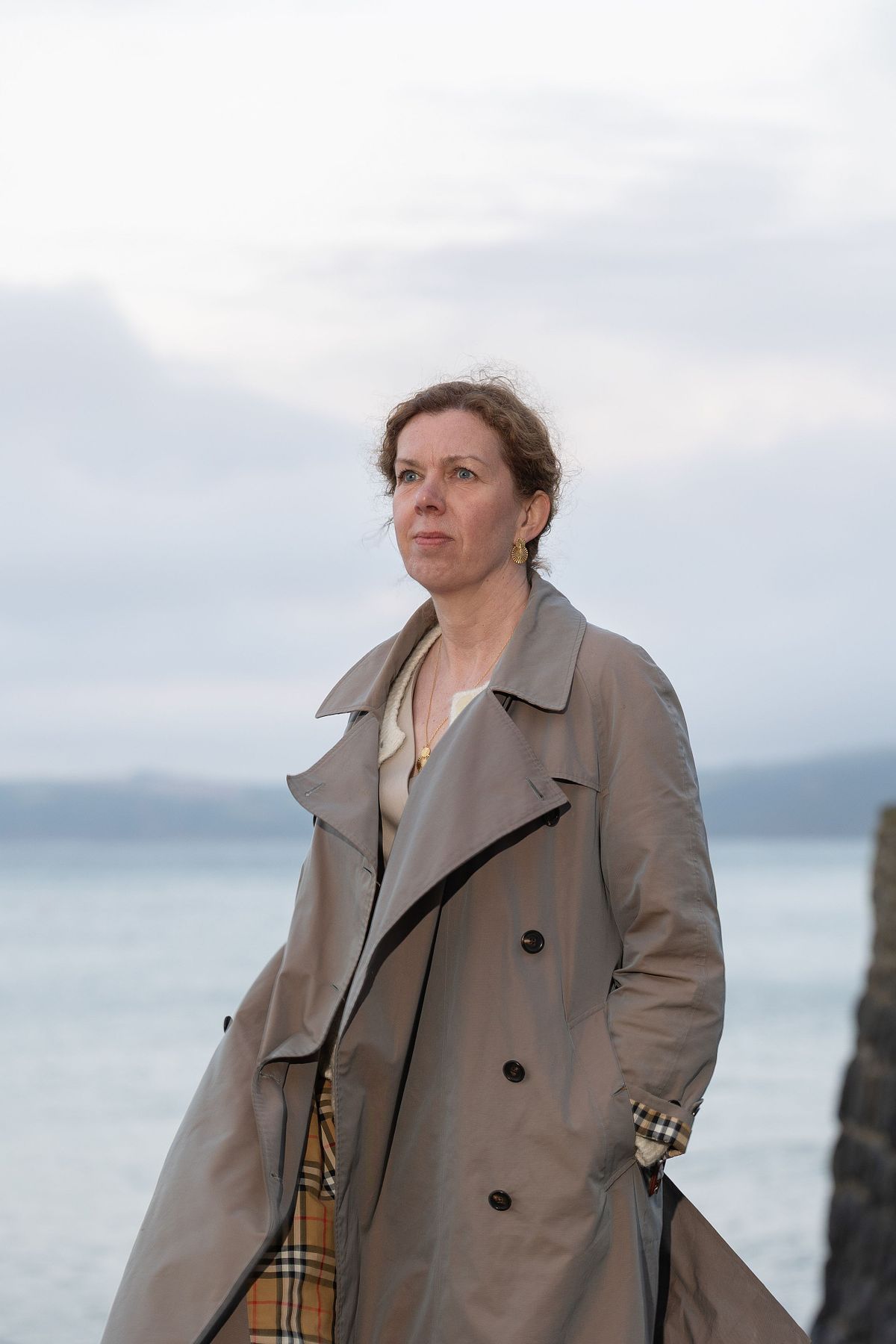 Kvinna i trenchcoat vid havet under molnig himmel, mjukt kv&auml;llsljus