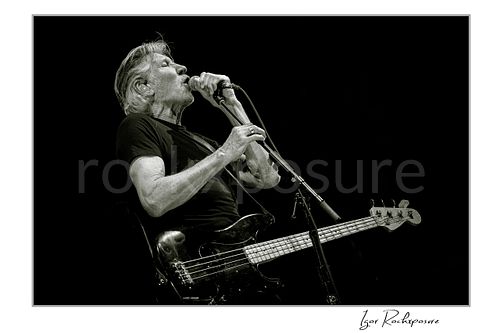 Horizontal black and white image of Roger Waters singing into a microphone while playing bass on stage, captured in a dramatic live performance against a dark background