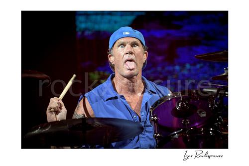 Horizontal color image of Chad Smith of Red Hot Chili Peppers at the drum kit in a tight live performance close-up, holding a drumstick with tongue out and vivid LED color behind him