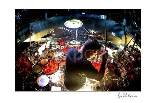 Horizontal color image of Korn drummer Ray Luzier seen from behind at the drums with one drumstick raised above the stage and crowd