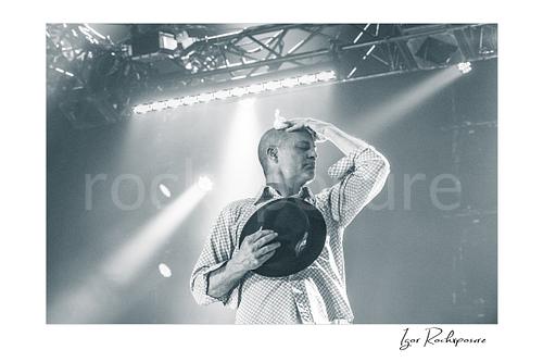 Horizontal black and white image of Gord Downie of The Tragically Hip in an emotional live performance moment, holding his hat to his chest while pressing a white cloth to his forehead under soft stage light