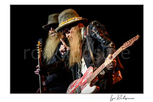 Horizontal color image of Billy Gibbons and Dusty Hill of ZZ Top performing live on stage, with Gibbons holding a red Telecaster and raising one finger while Hill stands behind him with bass in hand