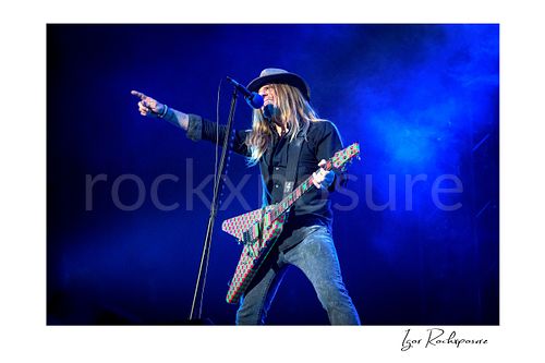 Horizontal color image of C.C. DeVille performing with electric guitar at the microphone during a Poison concert, pointing outward under blue stage light