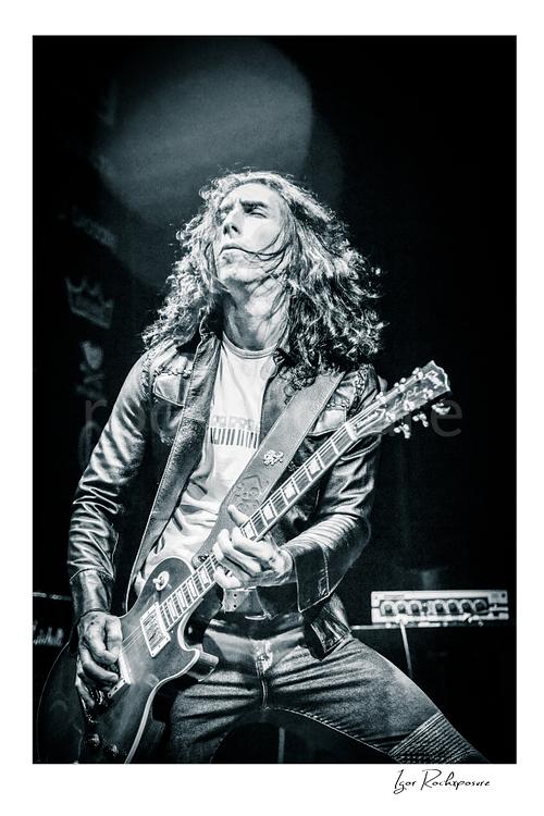 Vertical black and white image of Brent Woods playing guitar on stage with long hair and dramatic overhead light