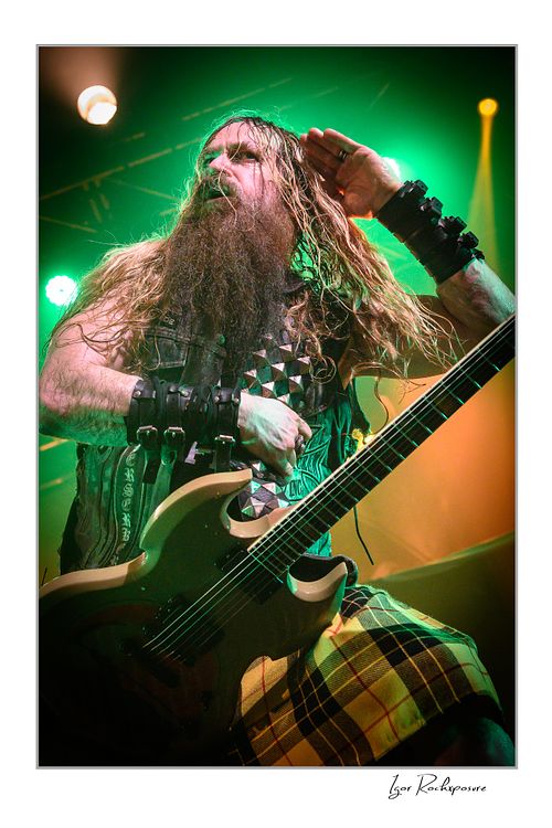 Vertical close-up of Zakk Wylde on stage under green lighting, wearing a denim vest and yellow plaid kilt, holding his guitar and saluting with his right hand.