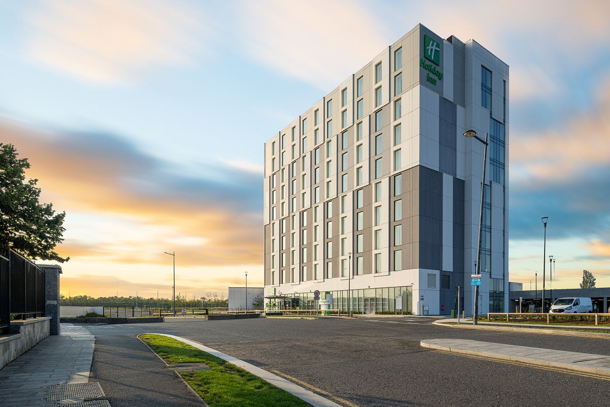 Exterior view of the Holiday Inn Dublin Airport, an IHG Hotel. The modern building features a multi-toned facade with rectangular windows. The Holiday Inn logo is visible. The image is taken at dusk, with a colorful sky.