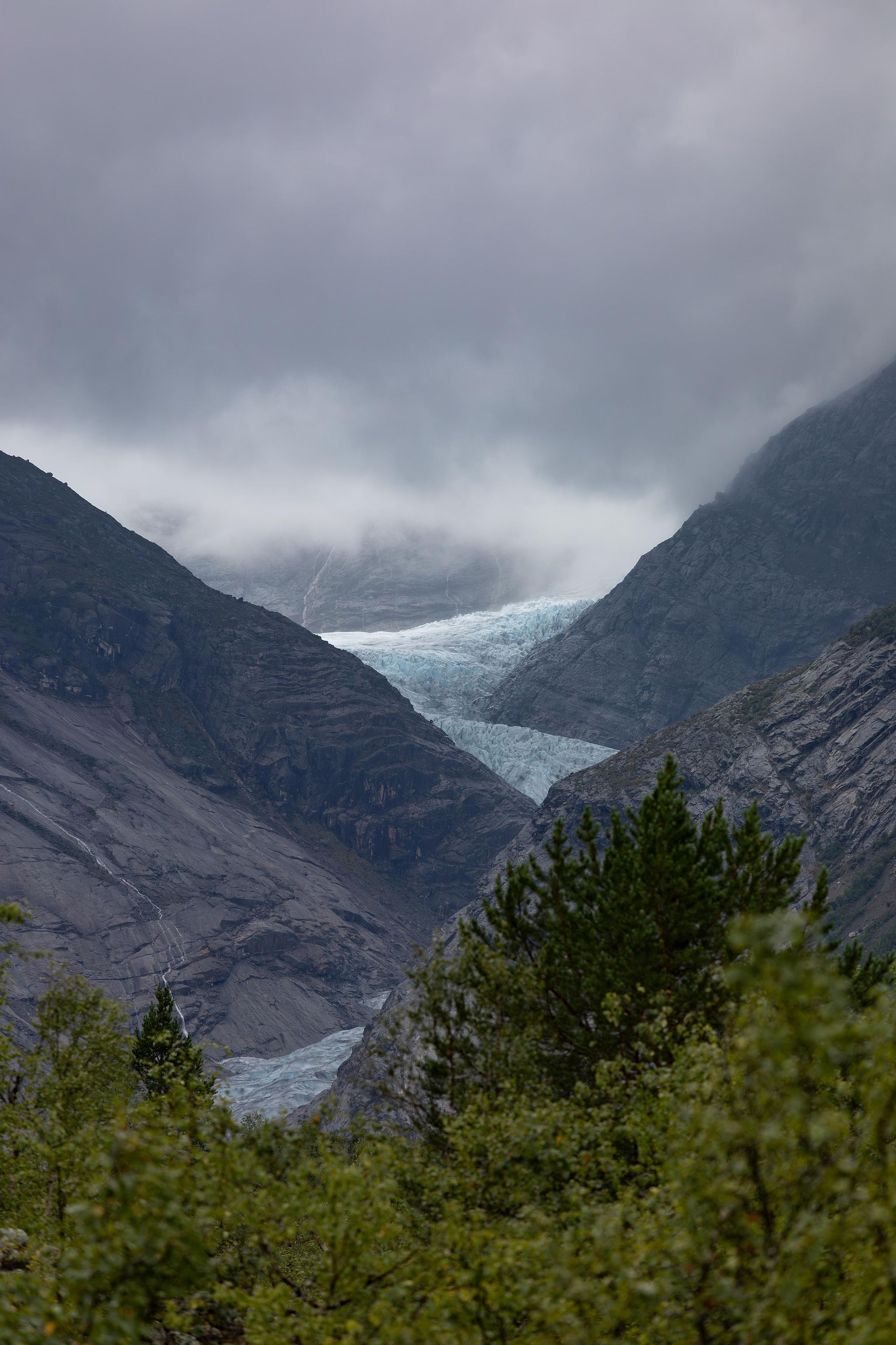 Glacier bleu encaissé entre deux montagnes rocheuses en Norvège, avec brume et ciel couvert en été.