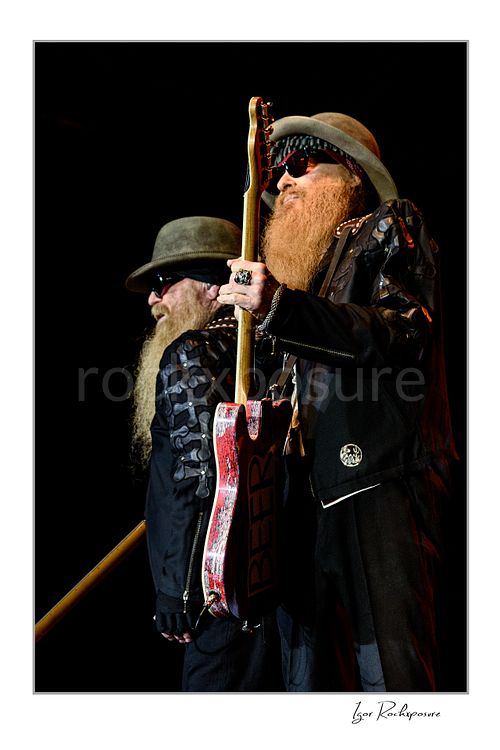 Vertical color image of Dusty Hill and Billy Gibbons of ZZ Top performing live on stage, standing side by side with signature hats, dark glasses, long beards, and guitars against a dark background