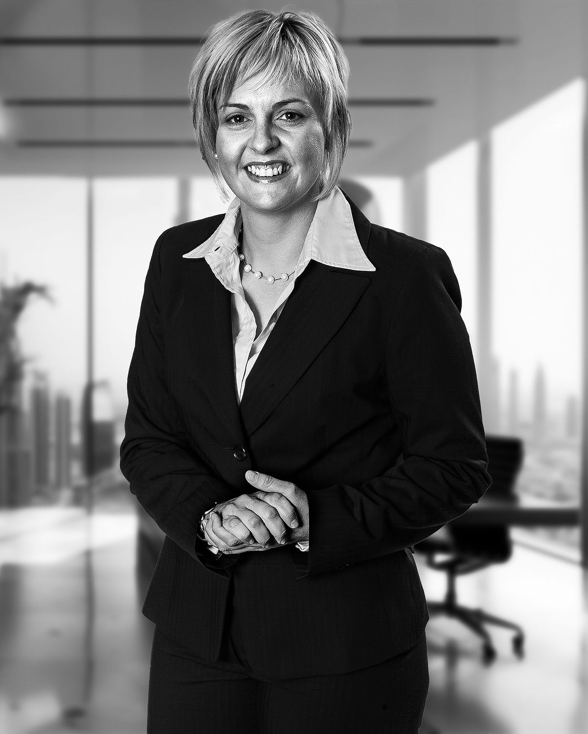 Corporate portrait of an Insurance business woman in her office. She wears a black suitm white shirt and tie. CIty in the background.