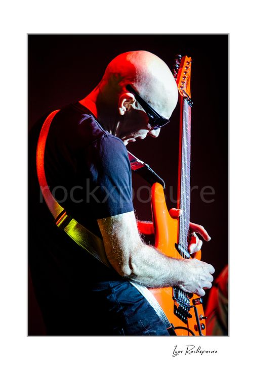 Vertical color image of Joe Satriani performing live with orange guitar, sunglasses, and dramatic red stage lighting