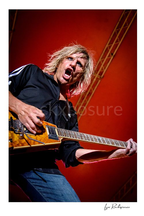 Vertical color image of C.C. DeVille performing with electric guitar during a Poison concert under vivid red stage light