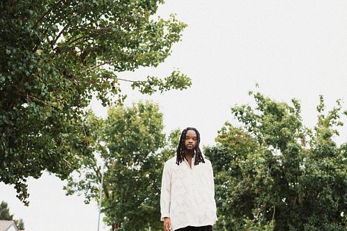 Musician Genesis Owusu stands outside with trees and white sky behind him while wearing a white long sleeve top, by Mila Filippone