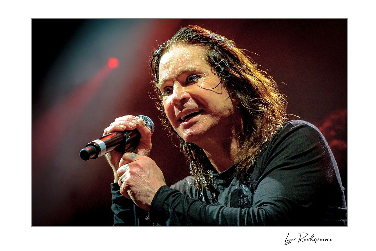Ozzy Osbourne of Black Sabbath singing into a handheld microphone during a live performance under warm red concert lighting