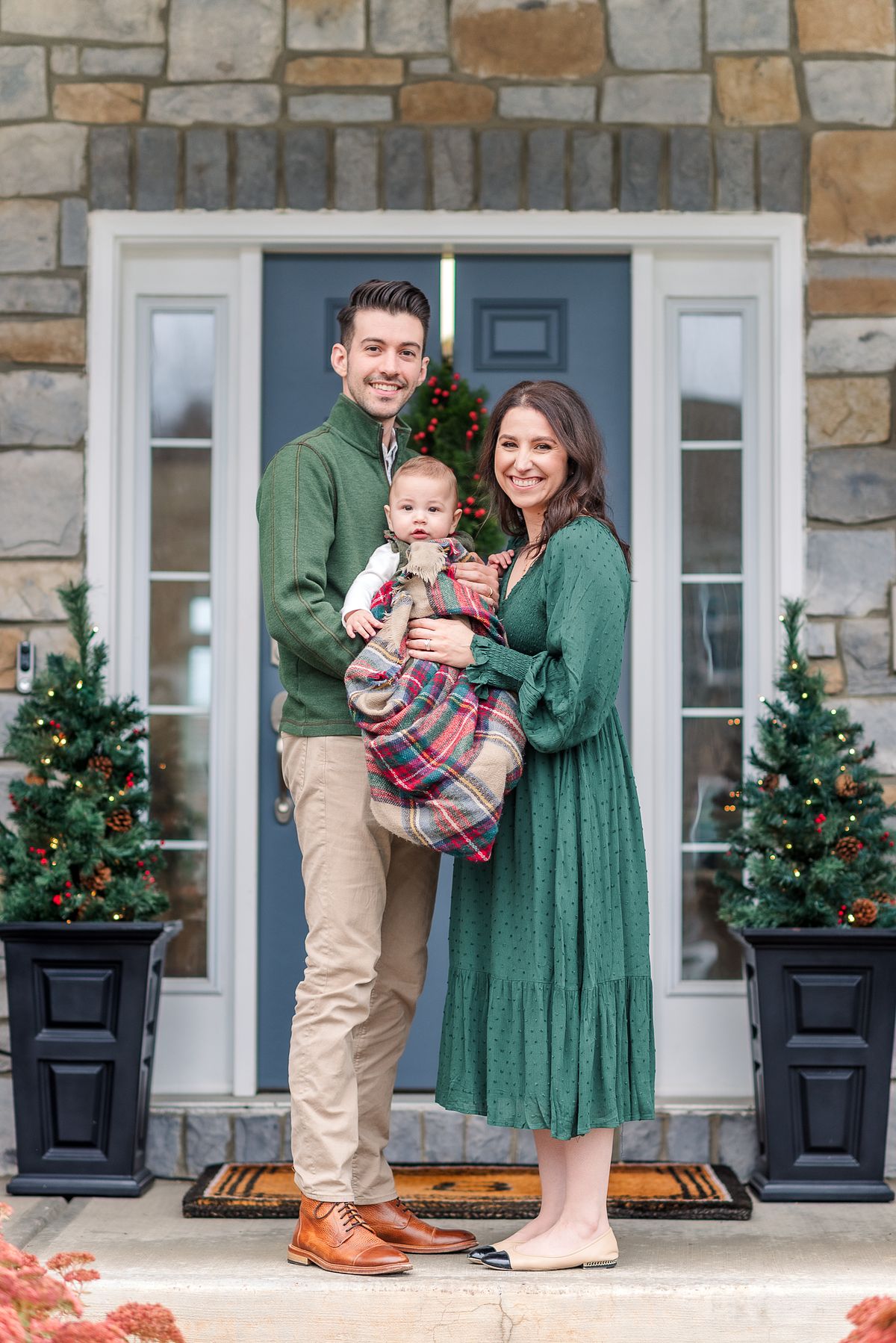 Husband and wife dressed in Christmas attire, holding their baby wrapped in a blanket on their decorated porch with Cranberry Township, PA newborn photographer