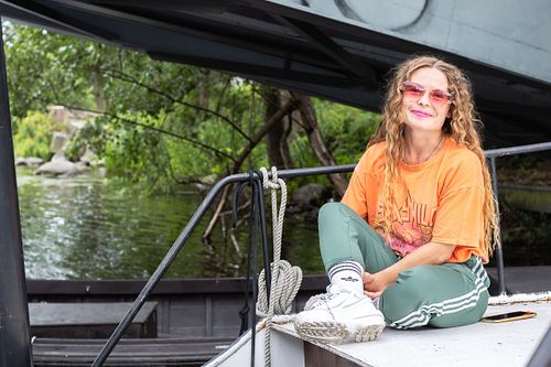 Lifestyle portrait of a smiling woman in orange T-shirt and green pants sitting on a boat, photographed in Stockholm by Mats Karlsson.