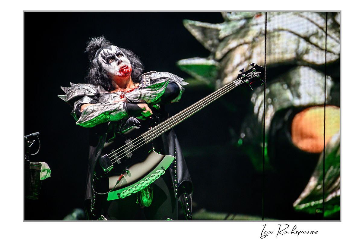 Gene Simmons of KISS in demon makeup with blood covering his mouth looking upward under green concert stage lighting during a dramatic stage moment