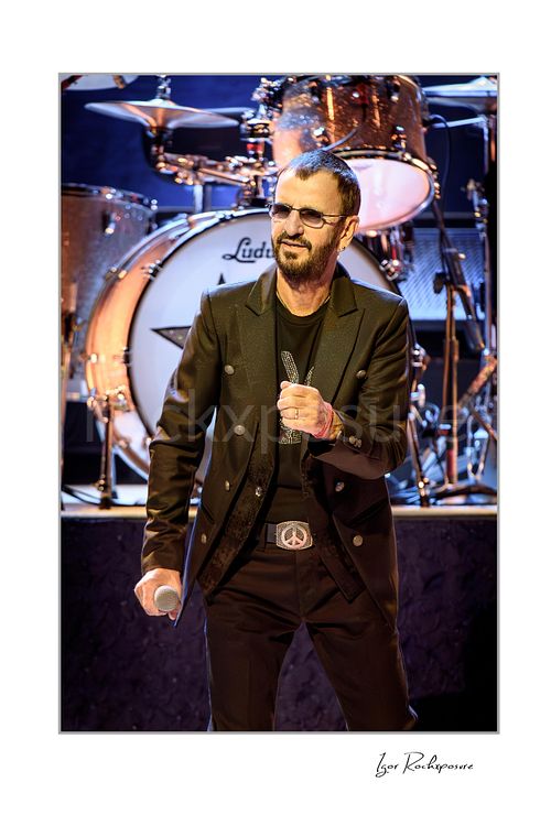 Vertical color image of Ringo Starr performing live on stage in a black jacket and sunglasses with a handheld microphone and drum kit behind him
