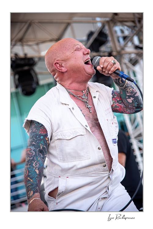 Vertical color image of Angry Anderson of Rose Tattoo singing into a microphone onstage in a sleeveless white outfit