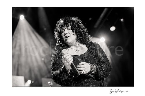 Horizontal black and white image of Ann Wilson singing into a microphone during a Heart concert in a dramatic close-up under bright stage lights