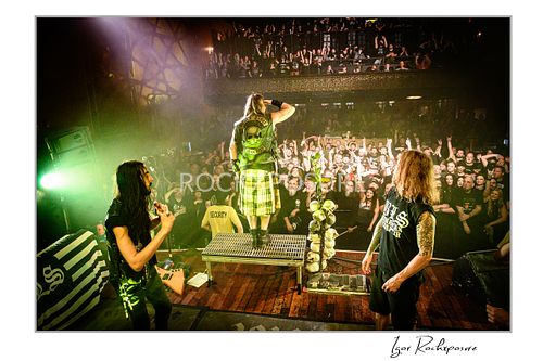 Wide shot from behind Zakk Wylde on stage with arms raised, cheering the crowd, flanked by John JD DeServio and Jeff Fabb under green stage lighting.