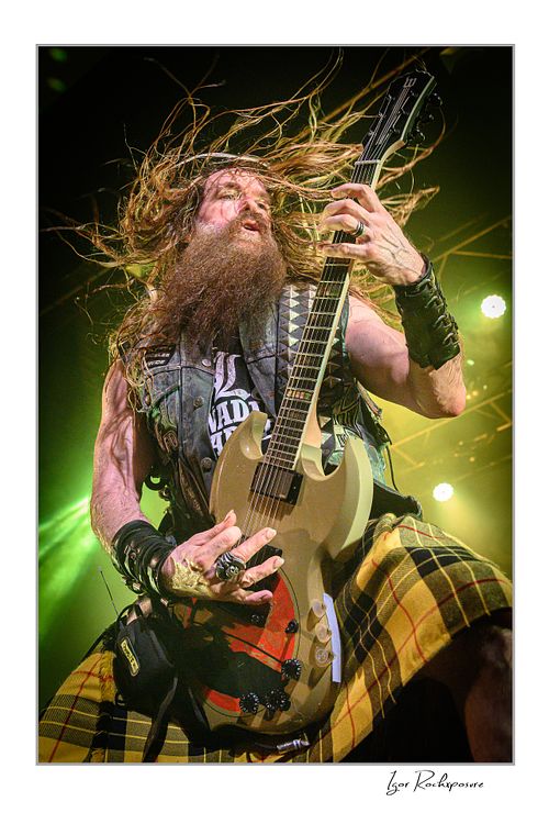 Vertical shot of Zakk Wylde playing guitar on stage, wearing a yellow plaid kilt and denim vest, with his head leaning back and long hair flying up under yellow and green lighting.