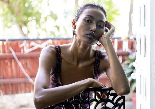 A portrait of an Indian woman in a purple top sitting on a chair on a balcony, with her arms placed on the backrest.