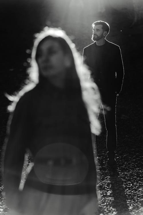 Ritratto in bianco e nero di donna che cammina verso la luce con il compagno sullo sfondo, strada nel bosco.