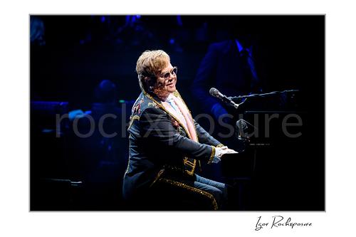 Horizontal color image of Elton John playing piano under deep blue stage lighting in a dark concert setting
