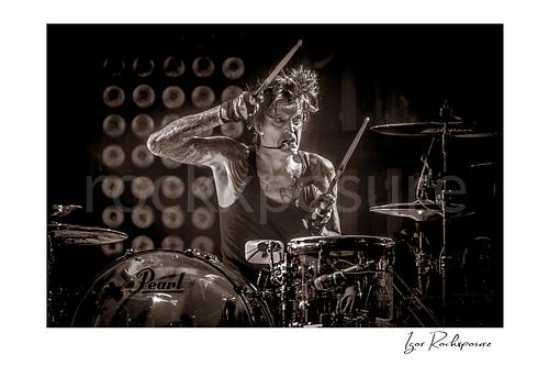Horizontal black and white image of Tommy Lee performing at the drum kit during a Mötley Crüe concert, holding raised drumsticks under dramatic stage light