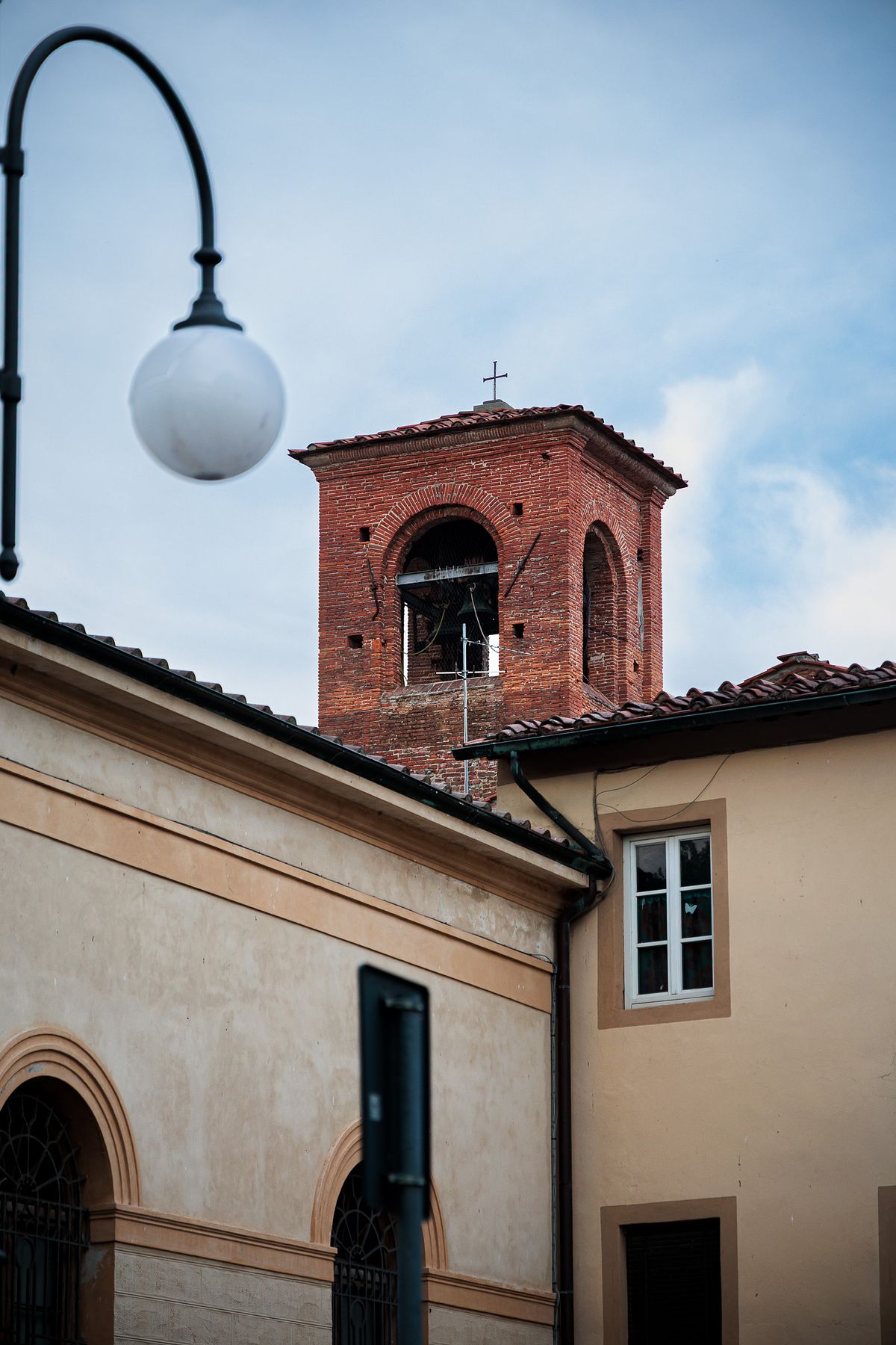 Street view of a red brick bell tower with a cross on top rising above light-colored buildings in Lucca, Italy, with a street lamp, sloped roofs and a partly cloudy sky creating a calm urban scene.