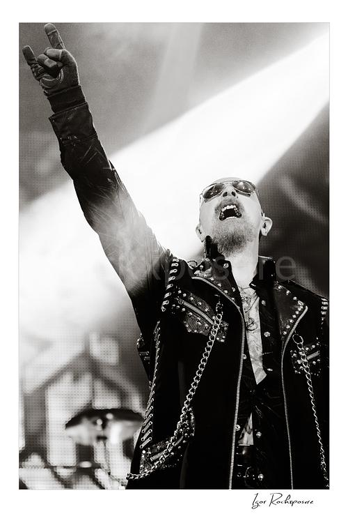Vertical black and white image of Rob Halford of Judas Priest performing live on stage, raising one arm while holding a microphone under dramatic concert lighting