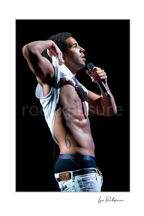 Vertical color image of Drake performing live on stage with a handheld microphone, lifted white shirt, and dark concert background