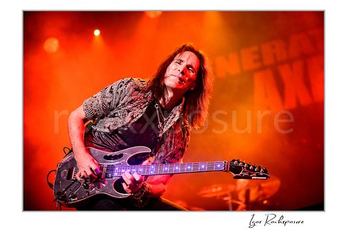 Horizontal color image of Steve Vai playing guitar under vivid orange stage lighting with glowing fret markers and light stage smoke