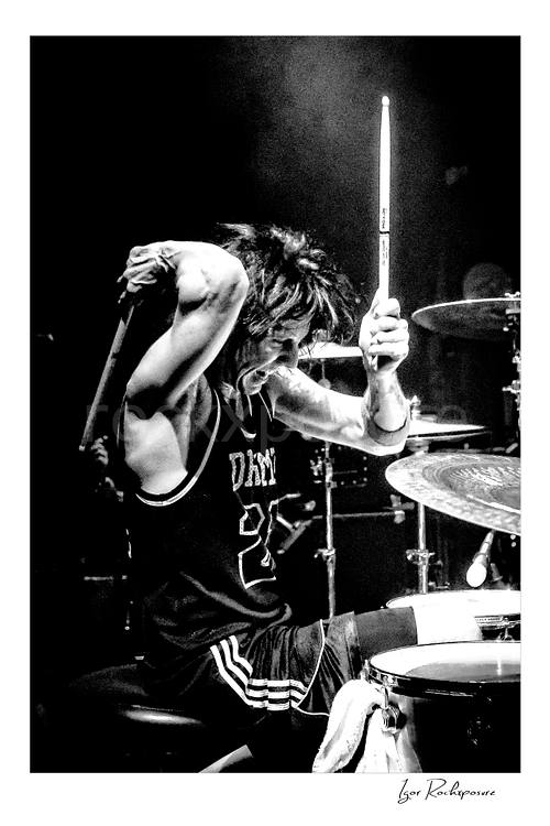 Vertical black and white image of Zoltan Chaney playing drums with raised drumsticks during a live performance