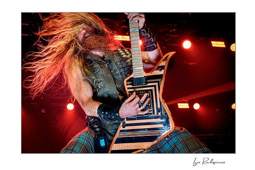 Zakk Wylde with hair in motion, playing guitar on stage under red lights.